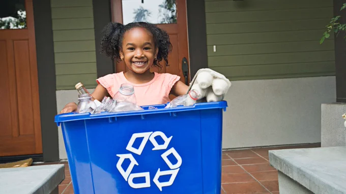 A child smiles while holding a recycling bin full of recyclable materials.