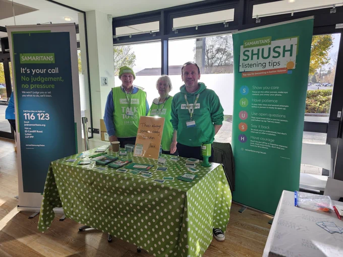 Three Samaritans volunteers standing next to a table with Samaritans promotional materials including leaflets, pens and pencils. There are two large banners that describe the support Samaritans offers on our telephone helpline. Call 116 123. We're available 24 hours a day. 