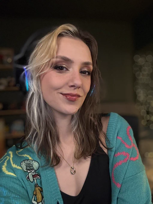 Ellie is smiling in a selfie in her stream room. She is wearing her shoulder length brown and blonde hair loose and wavy. She is wearing a black tank top under a teal SpongeBob cardigan and a black over ear headset