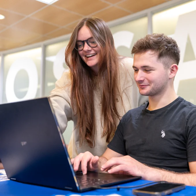 One person stands next to a person seated typing on a laptop
