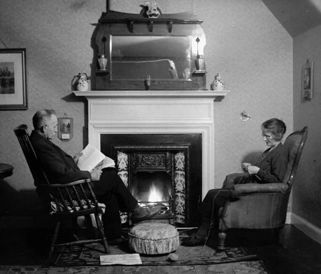 A fireplace with a burning fire and a mirror placed above the mantelpiece. A man sits on the left, reading. A footstool is beside his feet. A woman sits on the right, knitting. 