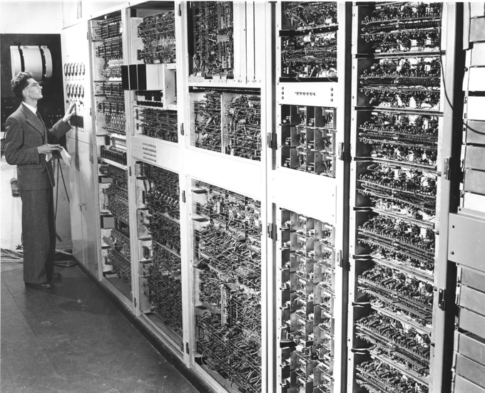 A black and white image of a man in a suit standing in front of the CSIRAC machine, a wall containing racks of wires, cables and buttons.