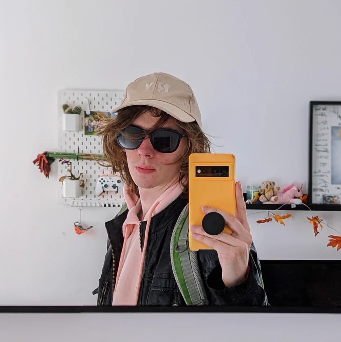 Topher taking a selfie in the mirror. They are wearing a pink hoodie with a leather jacket. Their phone is in a bright orange case.
They are wearing sunglasses and a light grey "NY" hat. Their hair is flowing around their face.

Behind them you can see a variety of trinkets - a custom Xbox controller, some flowers, a small teddy bear, a Rare branded candle.