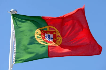 A photograph of the Portuguese flag against a blue background. It is a vertically divided green-red national flag with a coat of arms centred on the line between the two colours. 