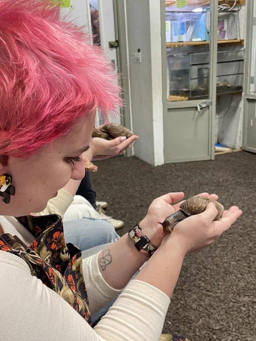 A person with a light skin tone and pink hair smiling and holding a snail.