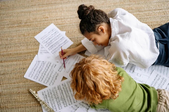 Two young people lying on a woven rug, surrounded by sheets of music. One person with dark hair in a bun is writing on a sheet with a pencil, while the other person with curly blonde hair watches. A flute rests on the rug near the sheet music.