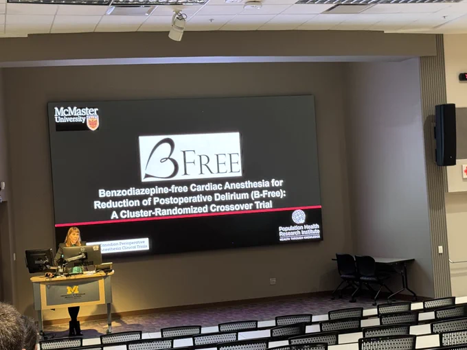 Female speaker at podium at front of auditorium. University of Michigan block M branded podium with computers on top. Slide reads “Benzodiazepine-free cardiac anesthesia for reduction of postoperative delirium (B-Free): A cluster randomized trial” McMaster Univeristy logo at top left. B-free logo in middle of screen features stylized heart in the shape of a letter B. “Canadian Perioperative clinical trials” logo at bottom left. “Population Health Research Institute: Health thru knowledge” logo at bottom right.