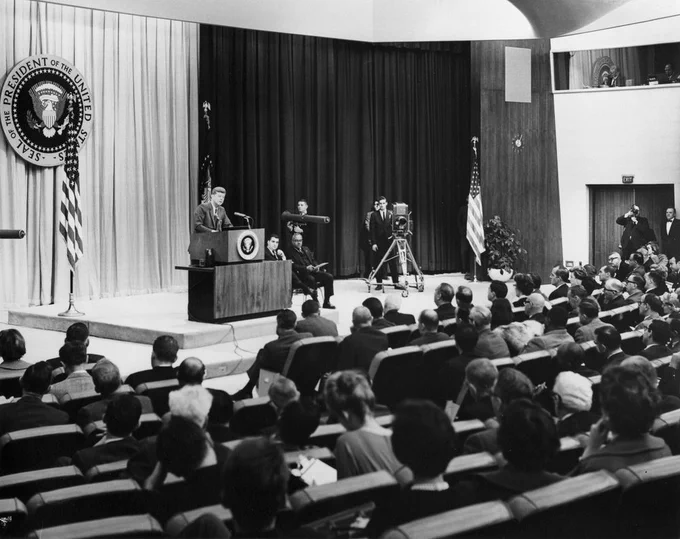 JFK stands a podium in an auditorium. His press secretaries are seated to his left on stage, and camera equipment is place behind them. Members of the press are seated in the auditorium.