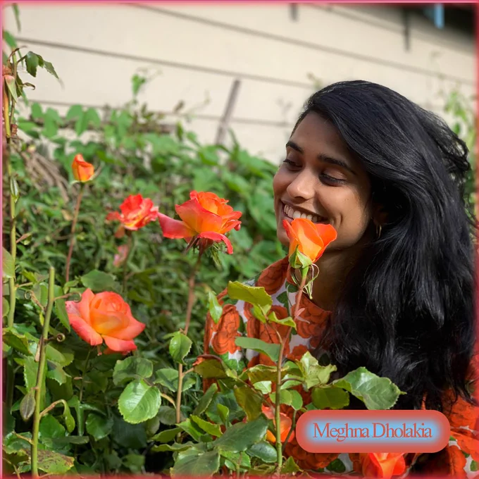 A brown skinned girl with long hair, leans into a coral rose bush. 