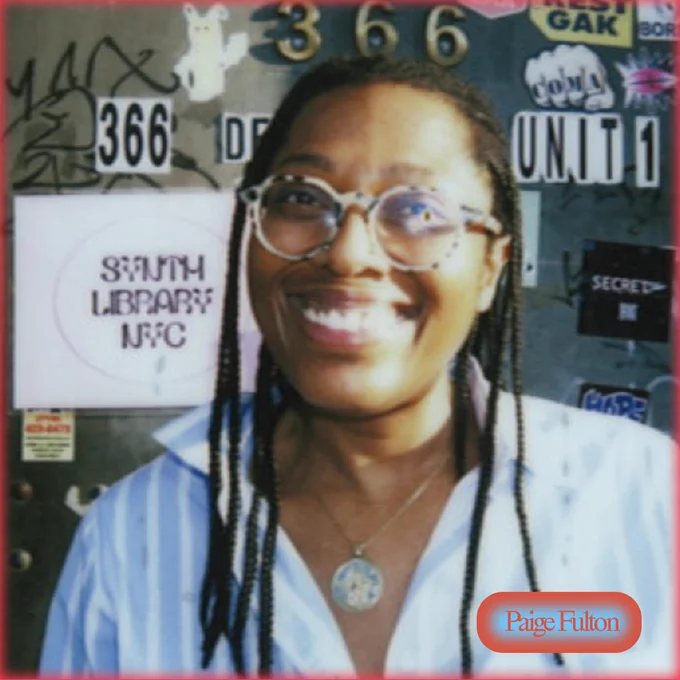 Paige, brown skinned black person smiling big with shoulder length braids and wearing a white and blue striped button down shirt, round tortoise glasses and a golden medallion chain. she's standing illuminated by the sun in front of a grafitti and sticker covered door.