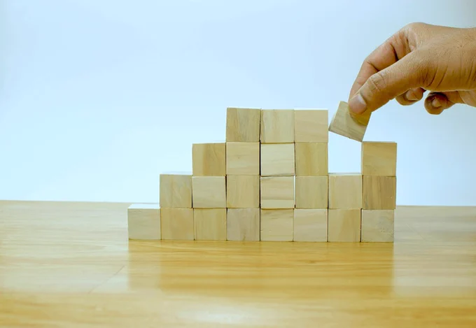 person putting a small wooden block in place on a stack of other wooden blocks (imagine-buddy-bTMTggEt5s4-unsplash)