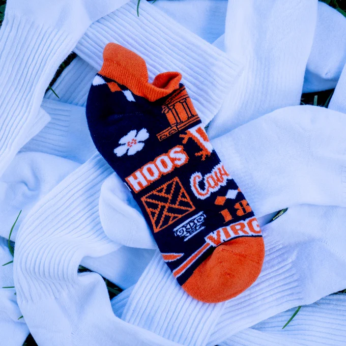 A University of Virginia-themed sock with orange and blue patterns, including text "Hoos Cav" and various UVA symbols, lying on top of plain white socks on grass.
