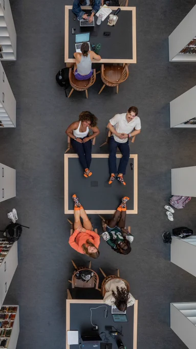 Overhead view of several individuals with feet up at desks in a modern library setting, with books and computers visible.