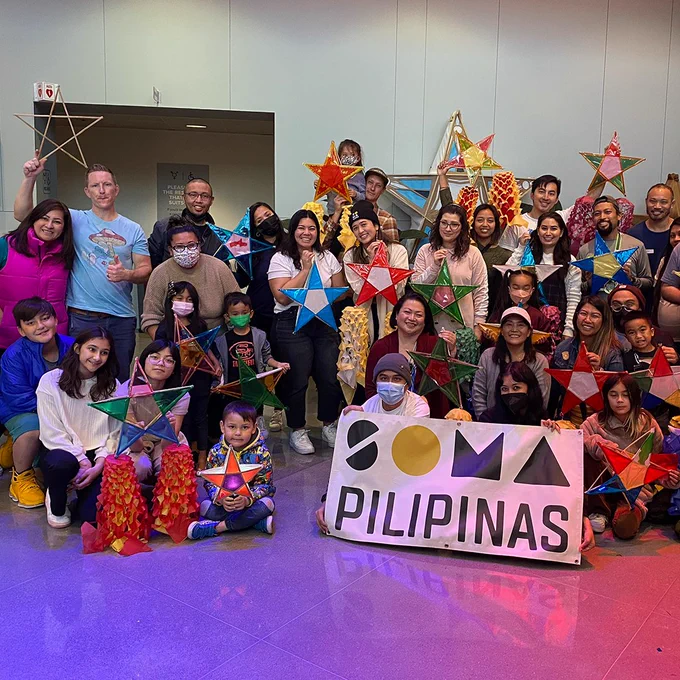 Group of people of various ages posing with colorful parol lanterns at a SOMA Pilipinas event in the YBCA lobby.