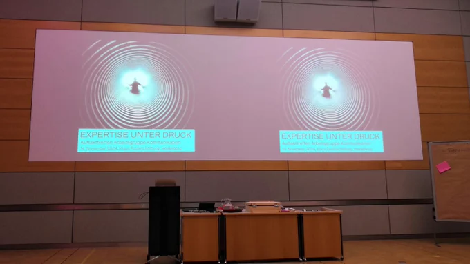 Eine Leinwand auf die das Titelbildes der Initiative "Expertise unter Druck" projiziert wurde. Darunter ein Rednerpult mit einem Medienkoffer.