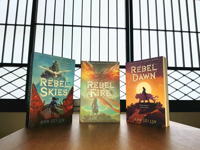 Three books lined up together in front of a screen window. The books are blue, green and orange respectively. Rebel Skies, Rebel Fire, and Rebel Dawn