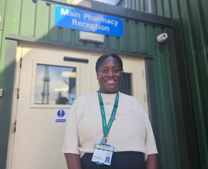A woman stands in front of a green building with a sign saying Main Pharmacy Reception behind her. She is smiling.