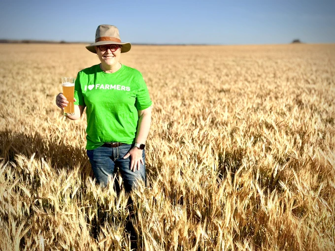 A woman with a brown hat, red round glasses, green shirt and denim jeans standing in a golden barley crop. She is holding a tall glass of non-alcoholic beer.