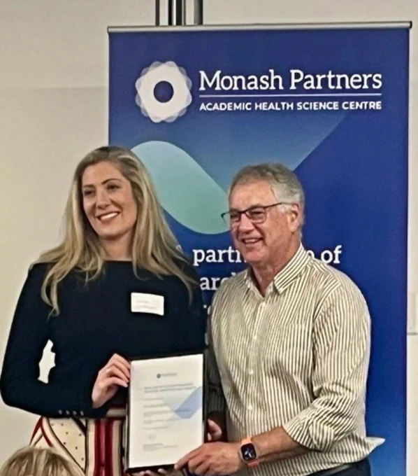 A woman with long blonde hair is shown standing with a man with glasses - they are holding a certificate and shown in front of a Monash Partners banner