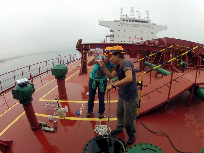Two scientists in hard hats stand on the red deck of a giant commercial ship, looking at water samples in a clear vial. 