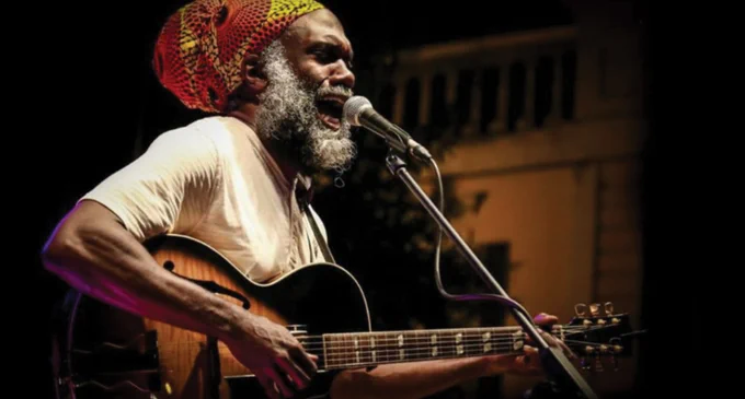 Musician Corey Harris performing live on stage with a guitar, wearing a colorful hat.