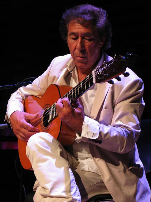 Photo of Juan Martín sat playing an acoustic guitar on stage, wearing a light-coloured suit and looking down at the guitar as he focuses on the music. 