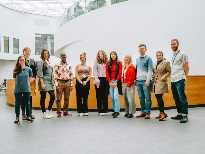 A photo of eleven people standing side by side, all looking at the camera. They're inside in the large atrium of a building, surrounded by white walls and in front of a long wooden desk