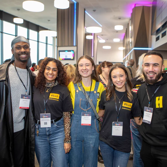 A group of five young adults standing together and smiling at a brightly lit event space. Two individuals wear black '#iwill Ambassador' t-shirts, and another is dressed in blue denim overalls with a yellow shirt. All are wearing name badges, with a diverse group of people visible in the background, chatting and engaging in the vibrant atmosphere.