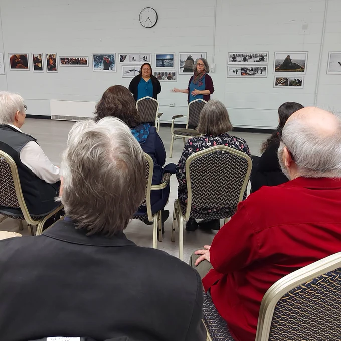 A photo of Camillie Fouillard, a white woamn with shoulder-length grey hair, introducing Annie, a round-faced Innu woman. They are standing in front of a seated crowd.