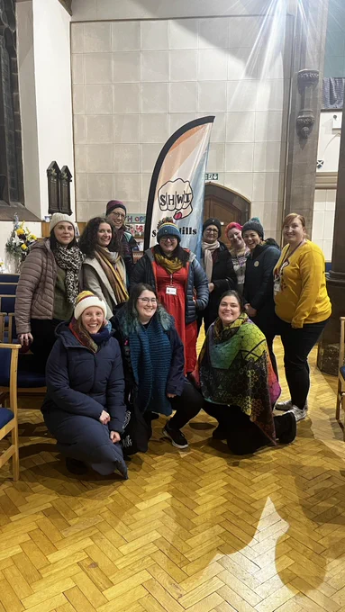 Members of the Seven Hills WI pose in front of the banner surrounding Chella. All are wearing hats and coats as it is cold inside the church meeting space.