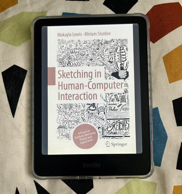 A Kindle device displaying the cover of the book Sketching in Human-Computer Interaction by Makayla Lewis and Miriam Sturdee. The cover features hand-drawn illustrations, sketches, and text surrounding the title. A red circle contains the subtitle: “A Practical Guide to Sketching Theory and Application.” The book is published by Springer. The Kindle rests on a patterned fabric background.