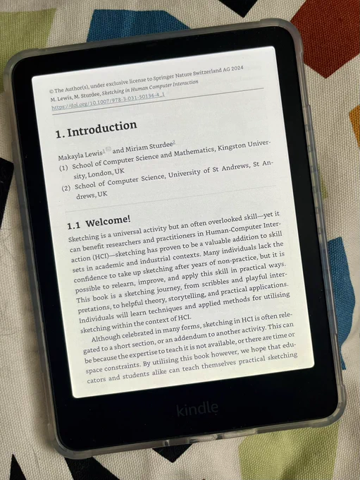 A Kindle device displaying the Introduction section of the book Sketching in Human-Computer Interaction by Makayla Lewis and Miriam Sturdee. The text includes the authors’ affiliations with Kingston University, London, UK, and the University of St Andrews, UK. The introduction highlights the value of sketching as a skill in Human-Computer Interaction (HCI) for researchers and practitioners, outlining the book’s focus on teaching sketching techniques and applications in HCI. The Kindle is placed on a patterned fabric background.