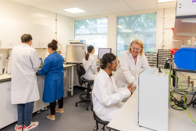 Five scientists in lab coats engaging in various tasks within a modern laboratory. Some are handling equipment and conducting experiments, while two are discussing research data joyfully.