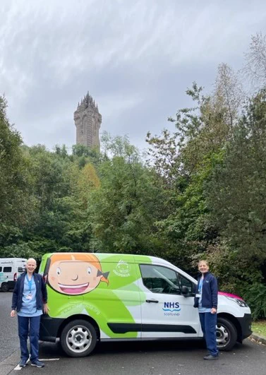 Alison and Yvonne standing beside the Childsmile van in a car park. In the background is the Wallace monument which is sitting behind some trees. 