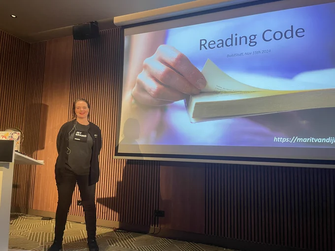 Marit standing near a big screen with the first slide of her talk Reading Code