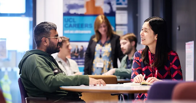 Five individuals engaging in a conversation at a UCL Careers event, with two pairs sitting at a table opposite each other, and one standing up. There are promotional banners in the background. 