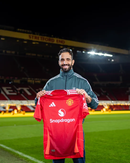 Ruben Amorim smiles while pitchside at Old Trafford holding a United home shirt up.