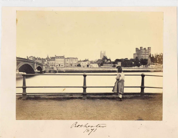 A sepia photograph. At the bottom it is captioned "Rochester 1871".  In the foreground is a railing separating dry land from the still River Medway. Stood near or possibly leaning on the railing is the figure of a young girl. We guess her age because her head is only a little higher than the railing. She is wearing simple period clothing, has shoulder-length dark hair and a dark, flat hat reminiscent of a sailor hat. The girl is looking to the left of the image, where the cast iron arches of the Victorian incarnation of Rochester Bridge is stretches across the river to the far bank. Along the far riverbank is a row of buildings of various shapes and sizes with lots of windows. Then we get to a wall and what looks like plants and trees. In the distance behind that is part of the tower of Rochester Cathedral minus its spire. Then further still, near the right edge of the picture is the stone shell of Rochester Castle. Above all of this is a pale, empty sky.