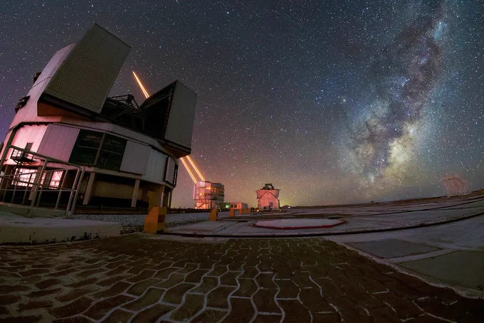 Nachtaufnahme von der Plattform des Cerro Paranal mit mehreren geöffneten Teleskopkuppeln , von einem der VLT-Hauptteleskope im Hintergrund gehen vier Laserstrahlen aus. Himmel rechts die Milchstraße