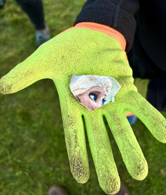 A photo of a child’s hand, wearing a bright yellow glove, holding a small fragment of plastic with a picture of Elsa from Frozen on it.