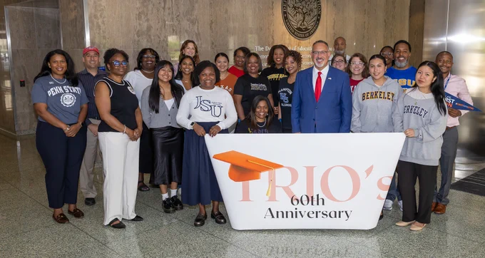 Secretary Cardona poses with a group of TRIO alumni and ED employees in front of a sign that reads "TRIO's 60th Anniversary"
