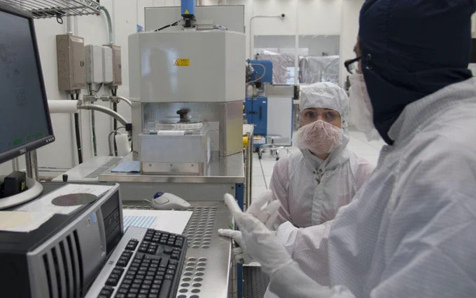Two people in full protective clean room gear talk in front of a computer and other scientific equipment. 