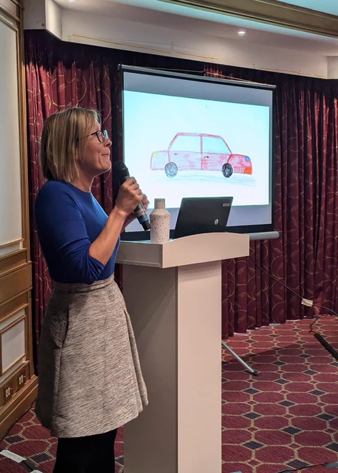 Laura stands at a podium in front of a drawing of a red car on screen.