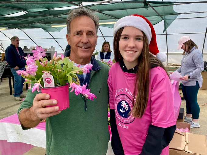 Two people standing and smiling at the camera. One is wearing a pink shirt and Santa hat and the other is wearing a green top and holding a pink flowering cactus in a pink pot.