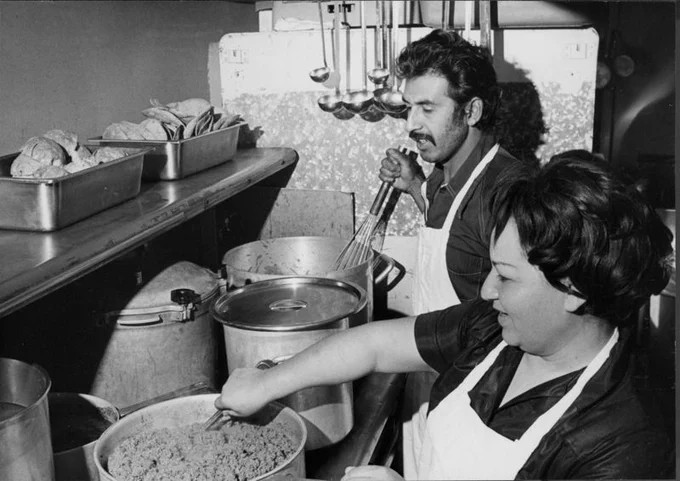 A black and white photo of a man and woman in a restaurant kitchen. The woman, in the foreground, is stirring a large pot of beans. 