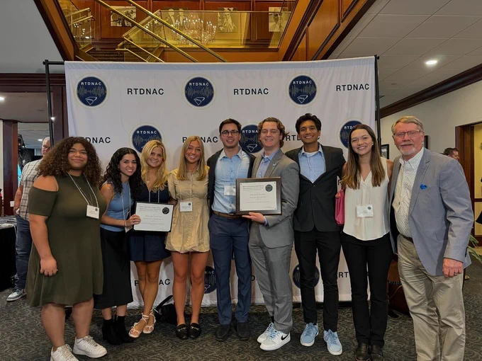 Nine people stand in front of RTDNAC step and repeat. Two hold awards.