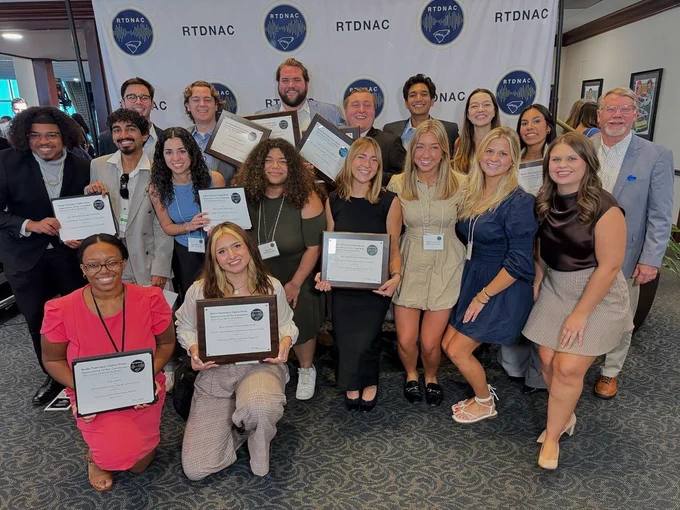 18 people stand in front of RTDNAC step and repeat. Many holding award plaques. 