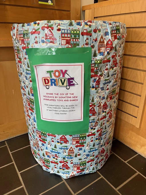 A barrel is wrapped with holiday paper featuring houses in the snow. A sign on front says, "Toy Drive, Share the joy of the holidays by donated new unwrapped toys and games!" 