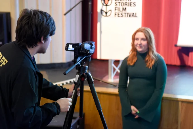 Person operating a camera on a tripod, focusing on another person standing by a banner of the "Earth Stories Film Festival.