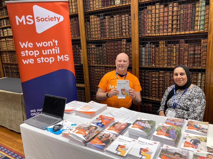 Image shows two people smiling to camera. The person on the left is wearing a bright orange T-shirt and is holding an information booklet about multiple sclerosis. The other person is smiling to camera, they are wearing a black and white patterned long sleeve top. They are also wearing a black heads scarf. Both people are sitting at a table with lots of information action booklets.